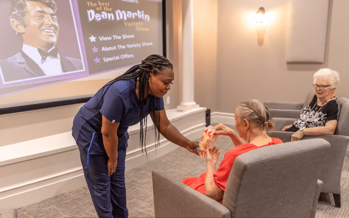 A caregiver in navy scrubs is handing a bag of popcorn to an elderly woman in a red shirt seated in a gray armchair. Another elderly woman with white hair and glasses sits in a similar armchair nearby. They are in a room with a large screen displaying information about 'The best of the Dean Martin Variety Show.'