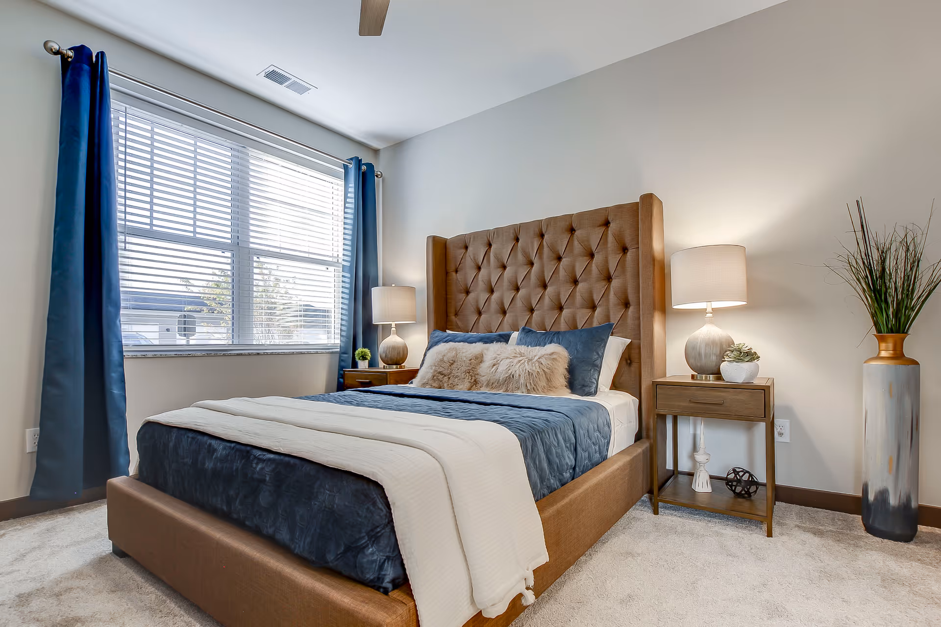A neatly made bed with a tufted brown headboard, blue and white bedding, and decorative pillows in a bright bedroom with a window covered by blue curtains. There are two wooden nightstands on either side of the bed, each with a lamp and decorative items. A tall vase with green plants is placed on the right side of the room.