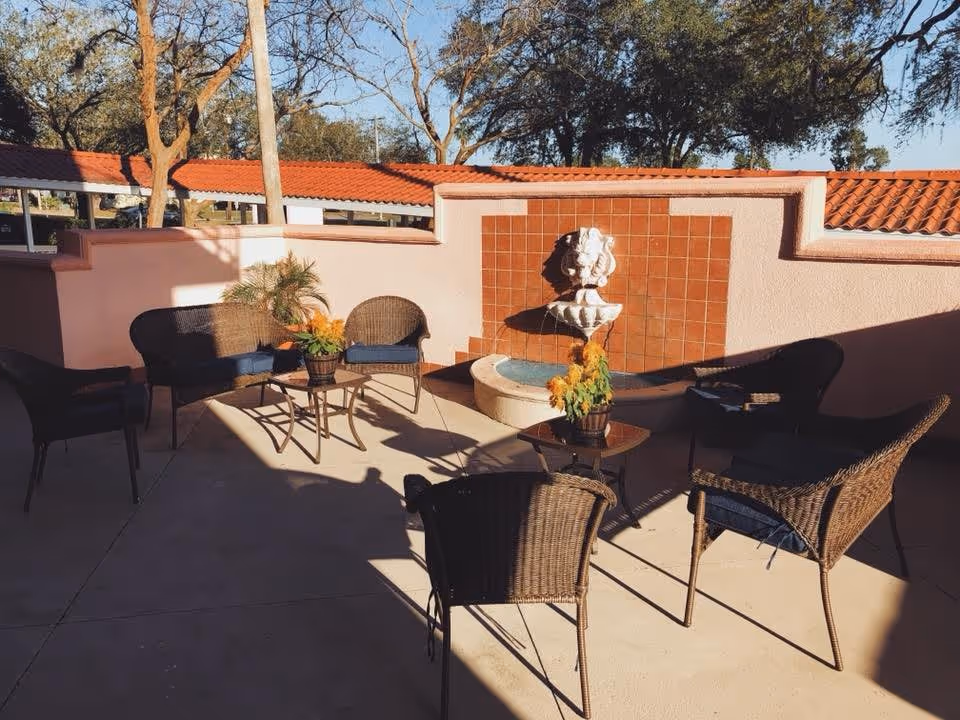A cozy outdoor patio at Osprey Manor featuring a fountain, wicker chairs, and potted plants under a clear blue sky.