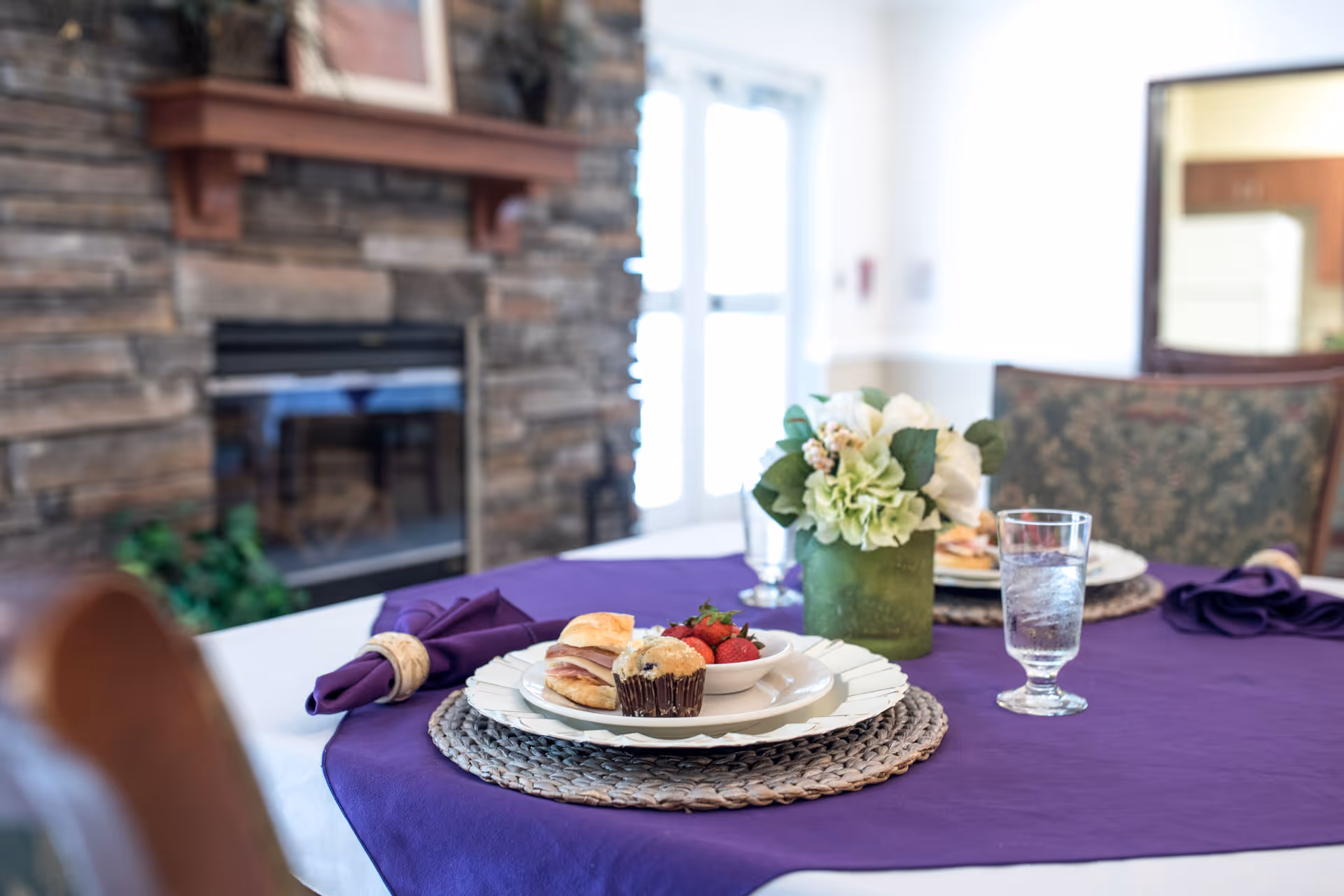 A dining table set with a purple tablecloth, white plates, a muffin, strawberries, a sandwich, a glass of water, and a green vase with white and green flowers. In the background, there is a stone fireplace and a door letting in natural light.