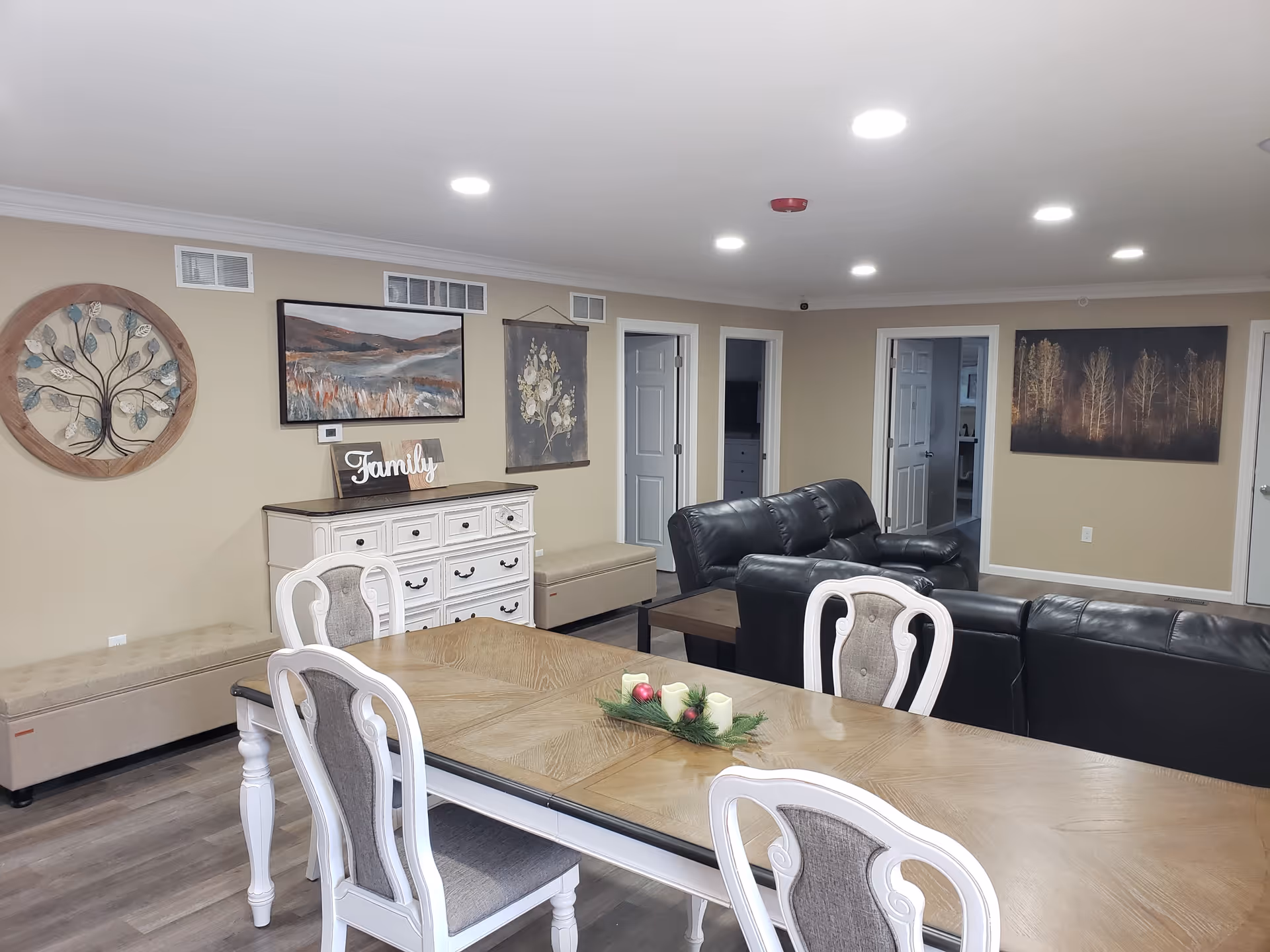 A cozy living and dining area with a wooden dining table and four upholstered chairs in the foreground. Behind the table, there are black leather sofas arranged around a coffee table. The walls are decorated with framed artwork and a decorative wooden tree piece. A white dresser with a 'Family' sign on top is also visible, along with beige benches along the walls. The room has recessed ceiling lights and light-colored walls and flooring.