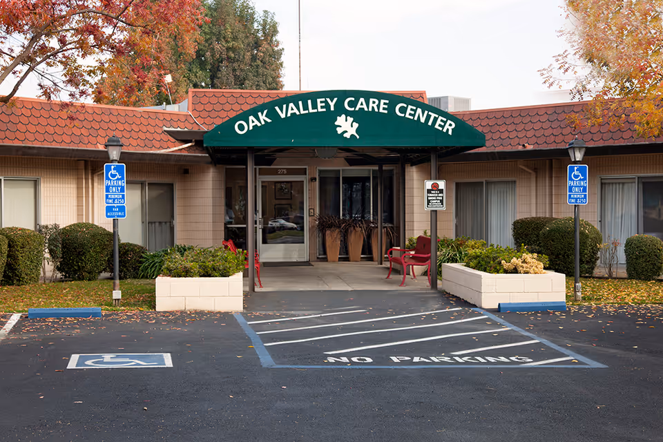Entrance of Oak Valley Care Center with a green awning displaying the facility name. There are two handicap parking spaces in front, marked with blue signs and painted symbols on the asphalt. The building has beige walls, windows with curtains, and some landscaping with bushes and planters. Red benches are placed near the entrance under the awning.