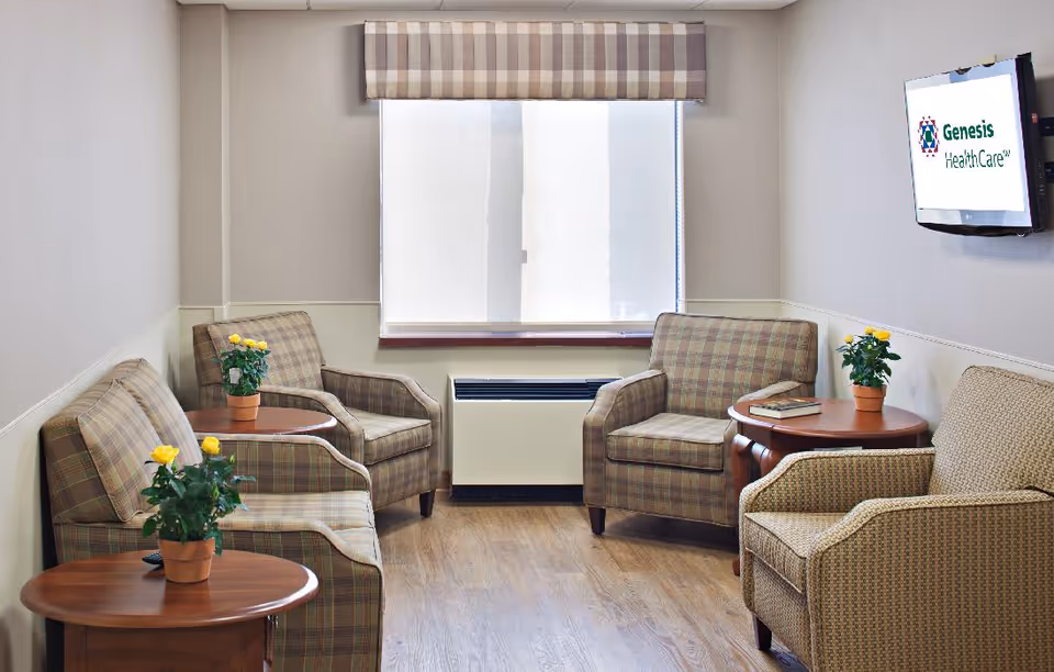 A small sitting area with four upholstered armchairs arranged around two wooden side tables, each holding a potted yellow rose plant. A window with a plaid valance is centered on the back wall, and a flat-screen TV mounted on the right wall displays the Genesis HealthCare logo.