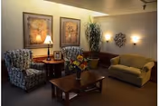 A cozy living room area with two patterned armchairs, a wooden coffee table with a flower vase, a small side table with a lamp, a large potted plant, a beige sofa, and framed artwork on the wall.