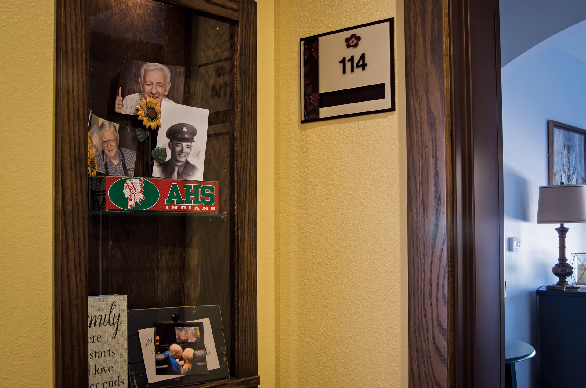 A hallway corner in a memory care facility showing a wooden display case with photos of elderly people and a military portrait, a sign with the room number 114 on a yellow wall, and part of a room with a lamp and framed artwork visible through an open door.