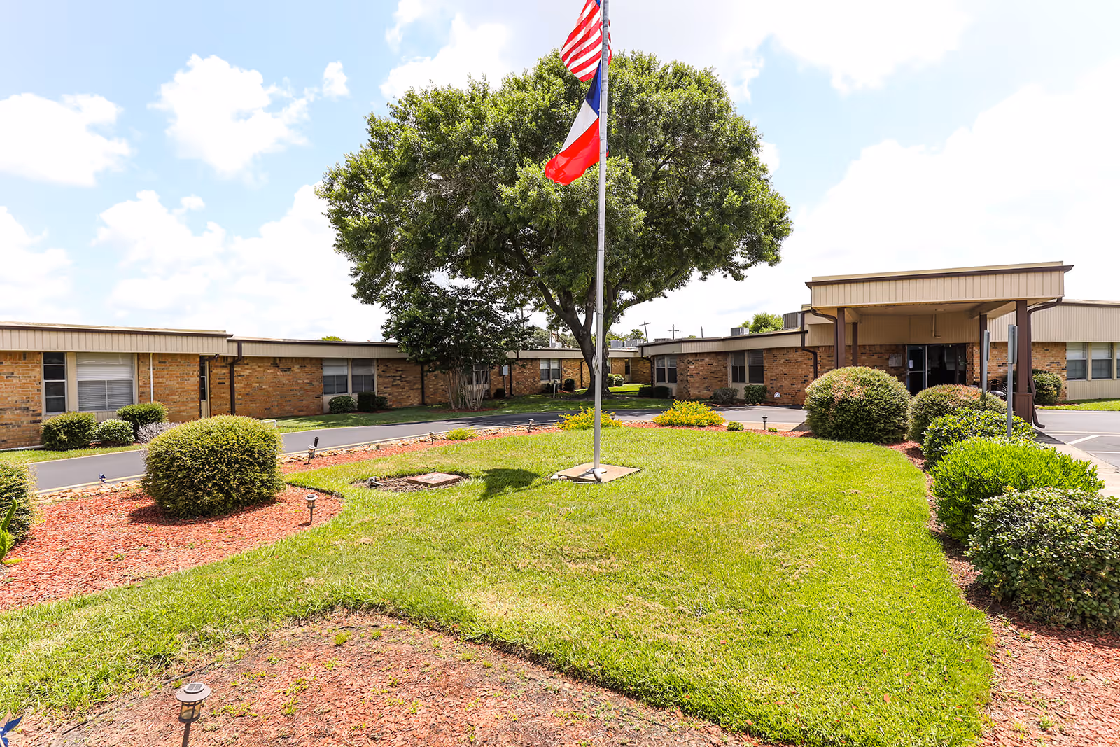 Exterior view of Brazos Healthcare Center showing a single-story brick building with a covered entrance, a well-maintained lawn, bushes, and a flagpole with the American and Texas flags in front under a partly cloudy sky.