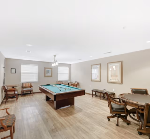 A well-lit recreational room with a pool table in the center, surrounded by chairs and small tables. The room has light-colored walls, wood flooring, two windows with blinds, framed artwork on the walls, and ceiling lights.
