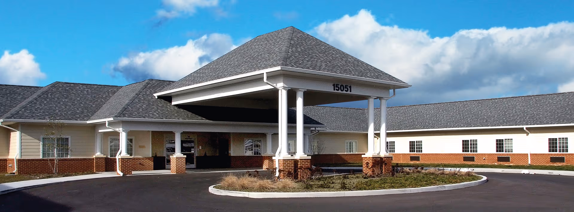 Front entrance of a single-story health and rehab facility with a covered porte-cochere, brick lower facade and a circular driveway under a blue sky.