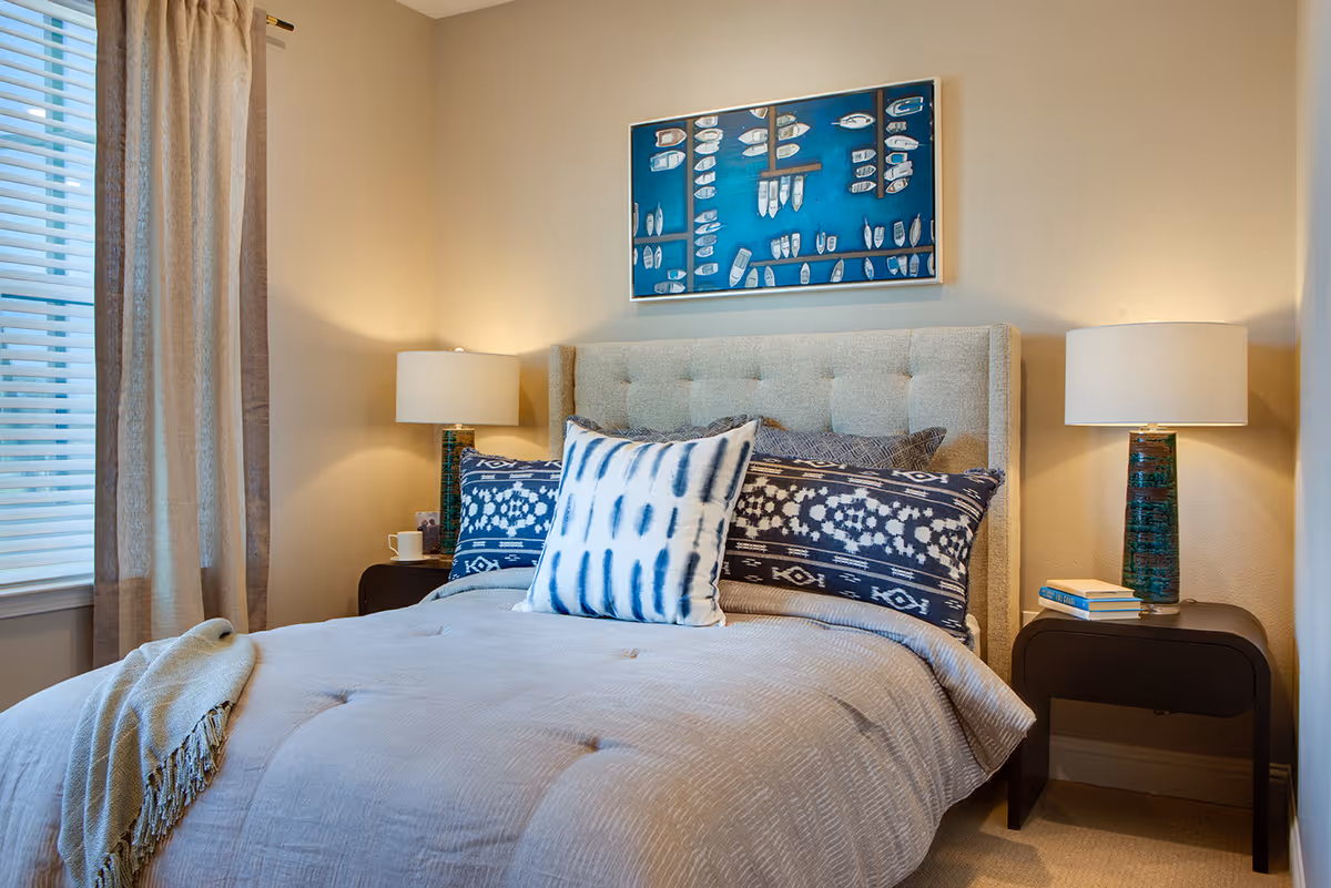 A cozy bedroom with a beige upholstered headboard, a bed made with a light gray comforter and decorative pillows in blue and white patterns. There are two dark wooden nightstands on either side of the bed, each with a lamp featuring a blue-green base and a white lampshade. A window with beige curtains is on the left side, and a framed artwork with a blue background and white shapes hangs above the bed.