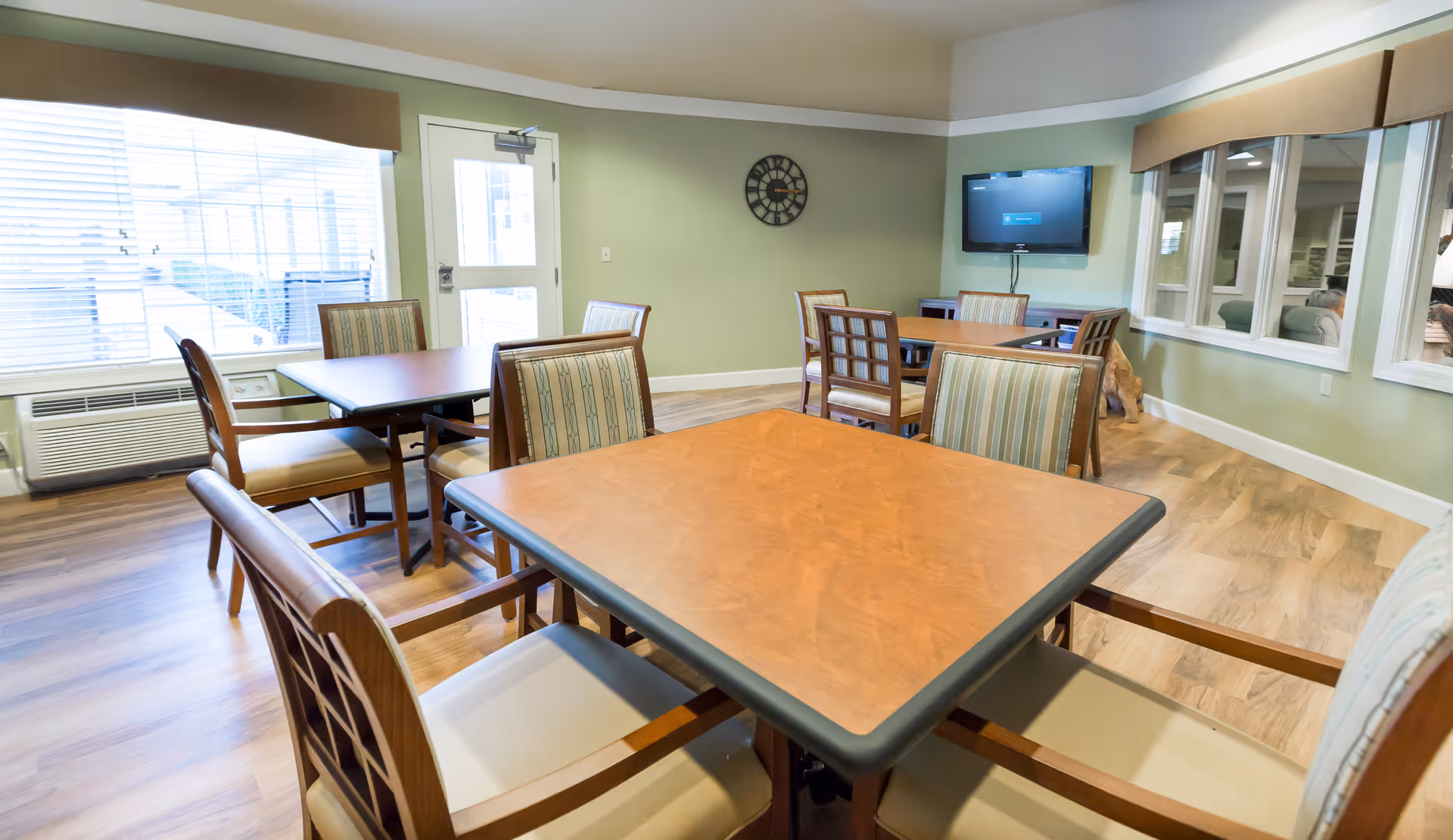 Communal dining room with square wooden tables and upholstered chairs, a wall-mounted TV, and large windows.
