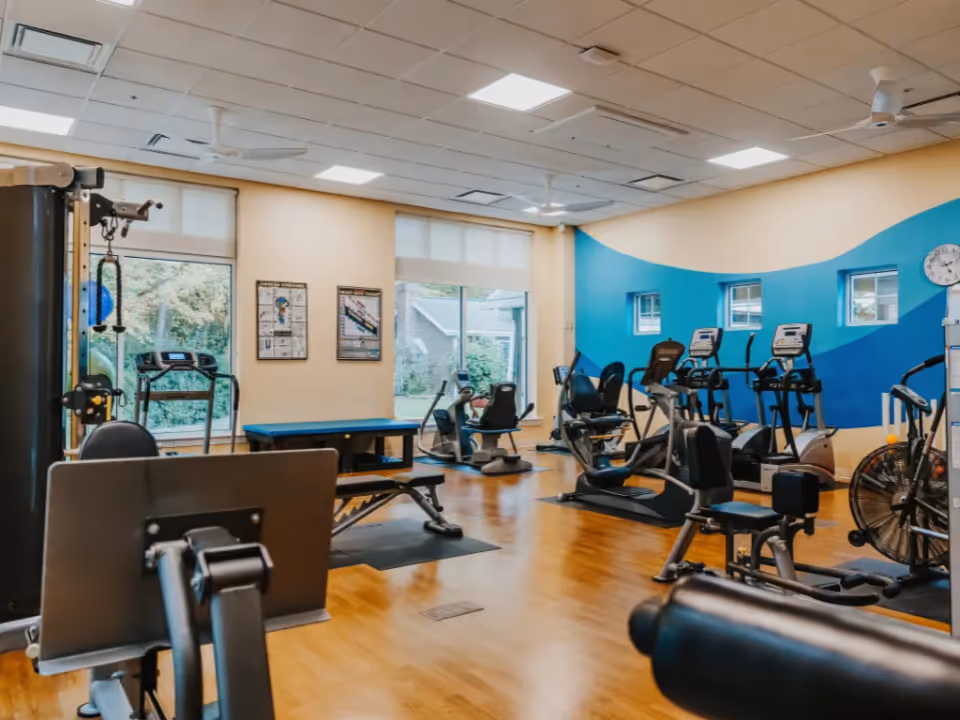 Bright fitness room with treadmills, stationary bikes, weight machines and benches on wood floors and large windows.