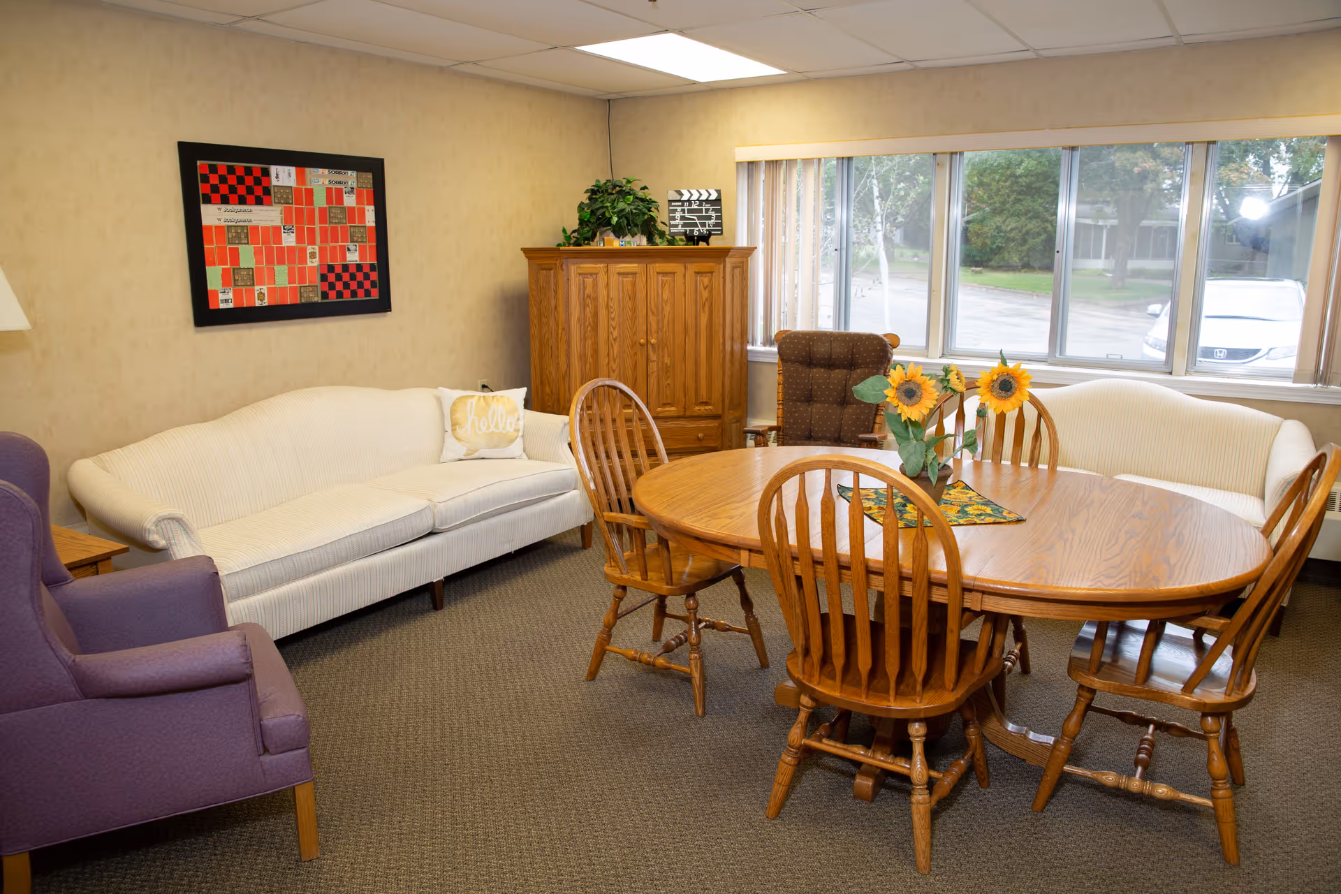 A cozy room with a wooden oval dining table surrounded by six wooden chairs. There are two white sofas and a purple armchair arranged around the table. A wooden cabinet with a plant and decorative items sits against the wall. A large window lets in natural light, and a colorful framed artwork hangs on the wall.
