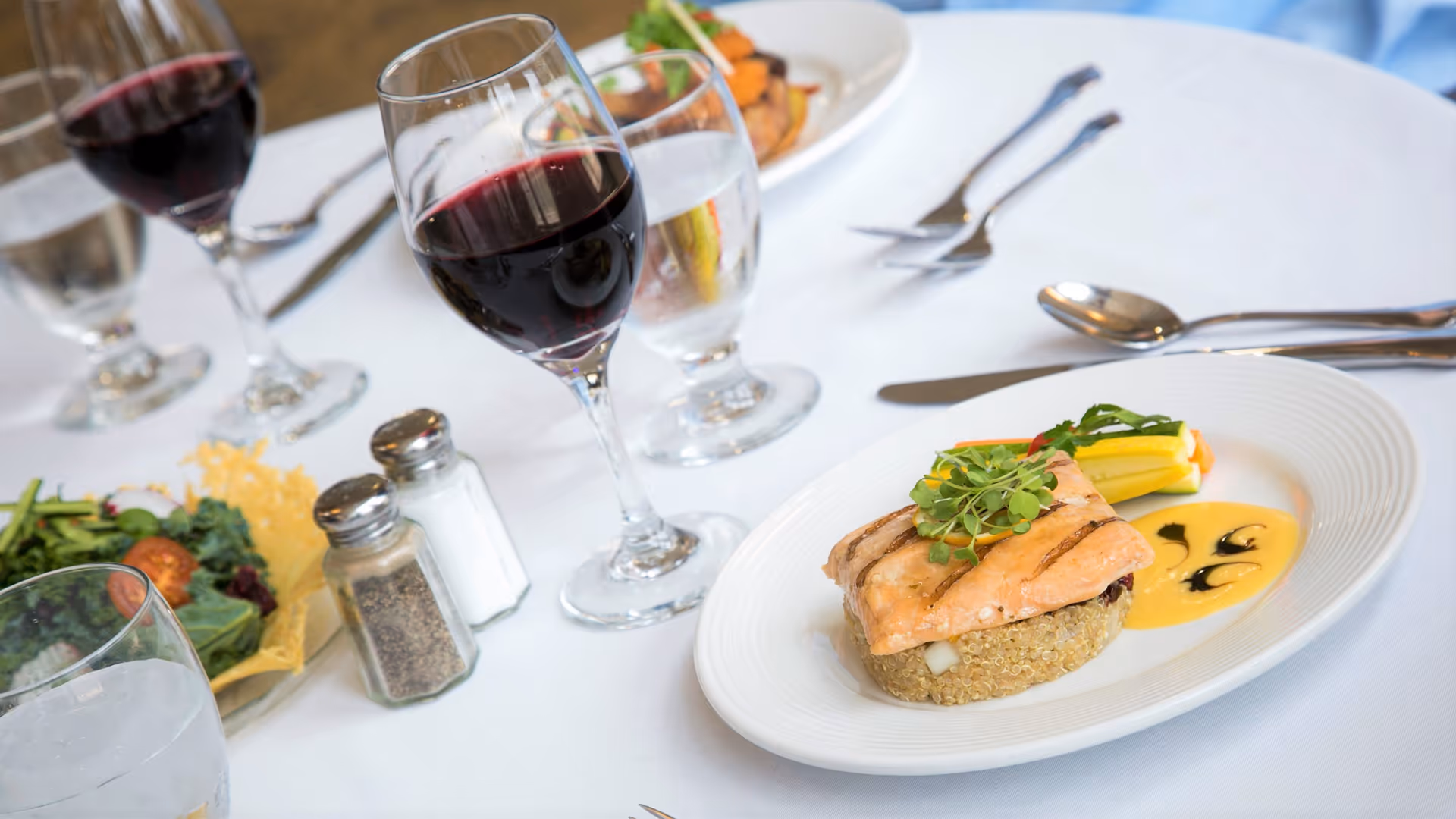 A fine dining table setting with a plated meal consisting of grilled salmon on a bed of quinoa, garnished with microgreens and served with a side of vegetables and sauce. The table also has glasses of red wine and water, a small salad, and salt and pepper shakers on a white tablecloth.