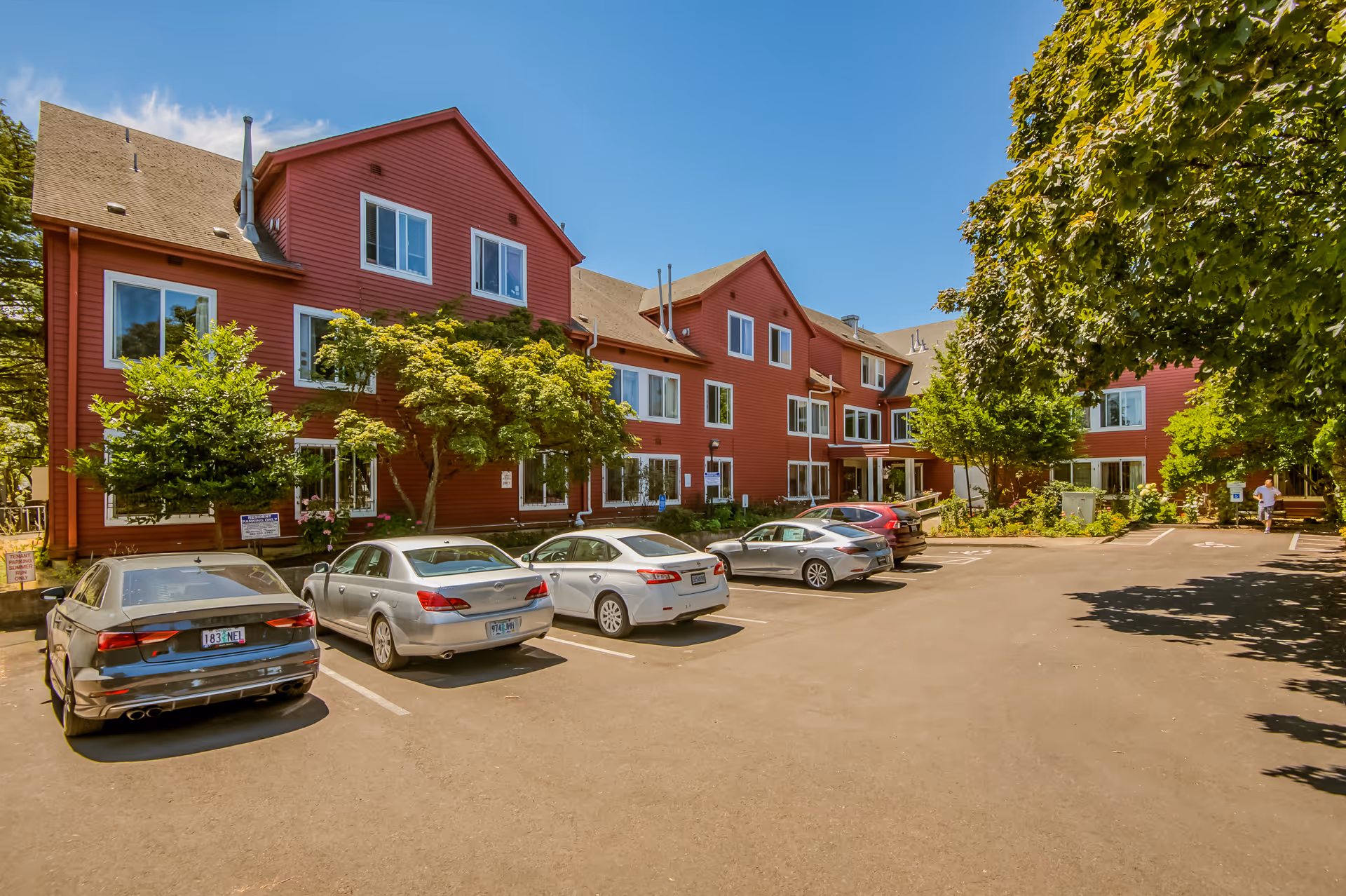 Exterior view of a red multi-story building with several windows, surrounded by trees and greenery. There is a parking lot in front with several parked cars under a clear blue sky.