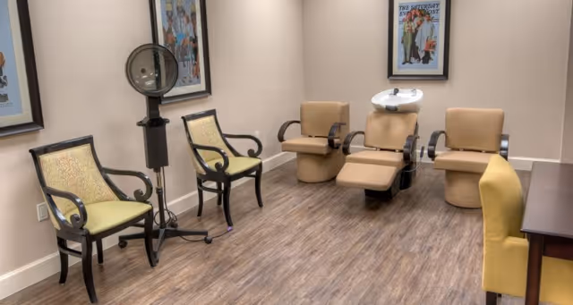 Interior view of a salon area in a senior living facility with two beige salon chairs with footrests and a hair washing station, two green cushioned chairs with dark wooden frames, a yellow chair, and framed artwork on the walls. The floor is wood-patterned.
