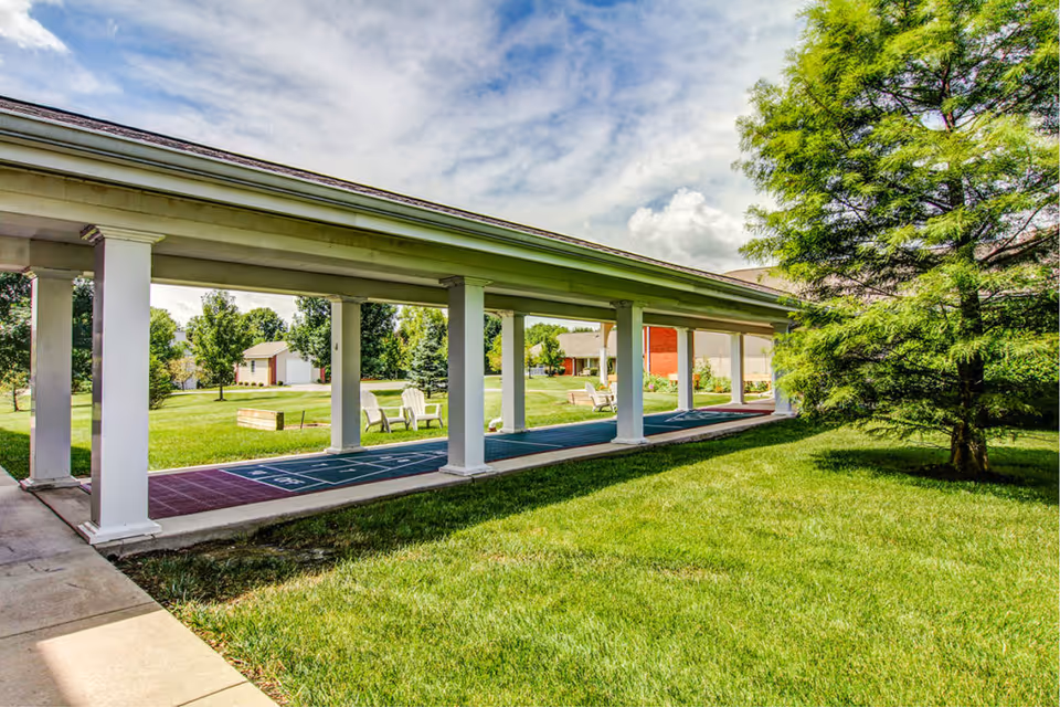 Covered outdoor shuffleboard court and walkway with white columns, lawn, and trees.