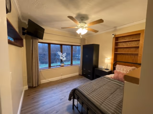 A cozy bedroom with a single bed featuring striped bedding and decorative pillows. There is a black wardrobe and a nightstand with a lamp beside the bed. A ceiling fan with lights is mounted on the ceiling. The room has a large window with beige curtains, showing an outdoor view with trees. A flat-screen TV is mounted on the wall opposite the bed.