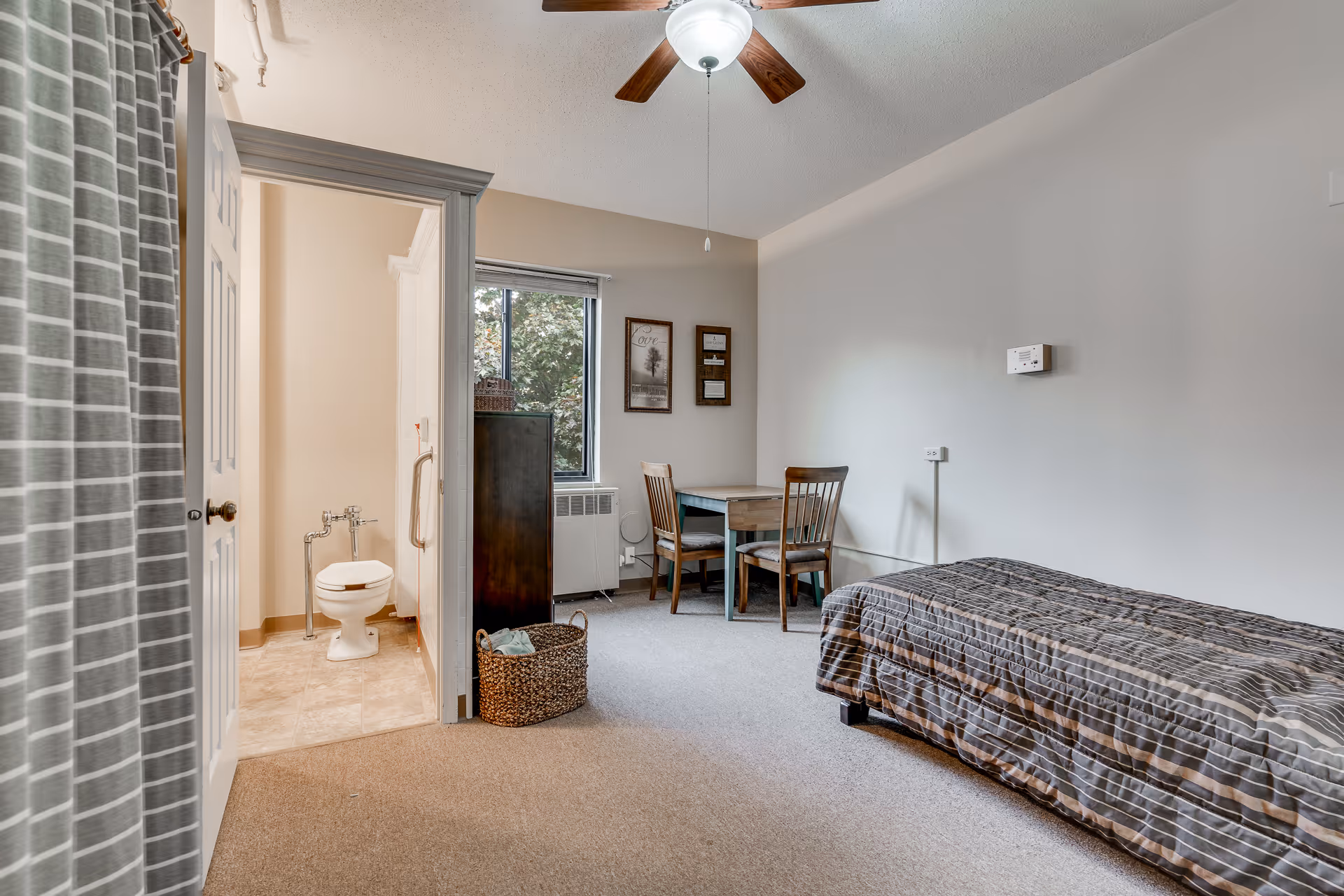 A senior living facility room with a single bed covered in a striped brown and gray bedspread, a small wooden table with two chairs near a window, a ceiling fan with light, and an open door leading to a bathroom with a toilet and grab bars. There is a basket on the floor and framed artwork on the wall.