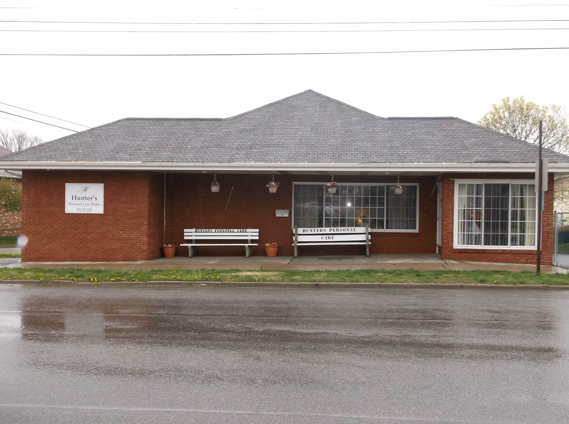 Front exterior view of Hunter's Personal Care Home, a single-story brick building with a gray shingled roof. There are two white benches with the text 'HUNTERS PERSONAL CARE' on the front porch, hanging flower pots above, and a large window to the right. A sign on the left side of the building reads 'Hunter's Personal Care Home' with a phone number.