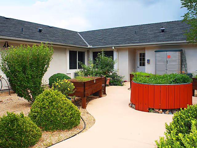 Outdoor courtyard area at Willow Wind Assisted Living featuring a paved walkway surrounded by green shrubs and raised garden beds with plants. The building exterior has beige walls and a dark shingled roof.