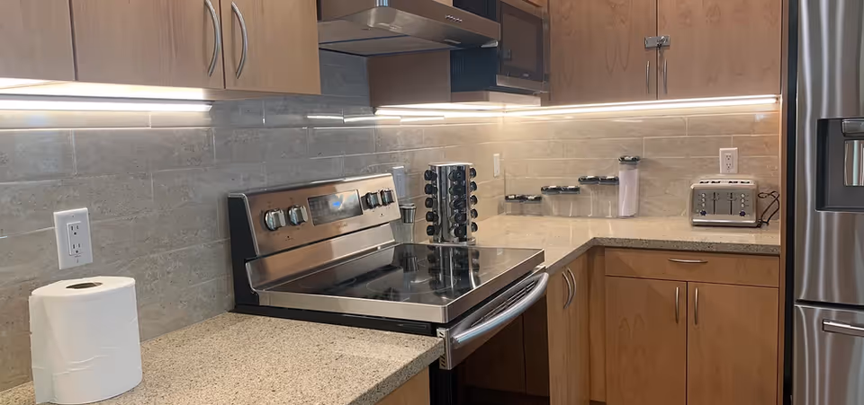 Bright modern kitchen with a stainless steel stove and refrigerator, light wood cabinets, under-cabinet lighting, toaster, spice rack, and a roll of paper towels on the countertop.