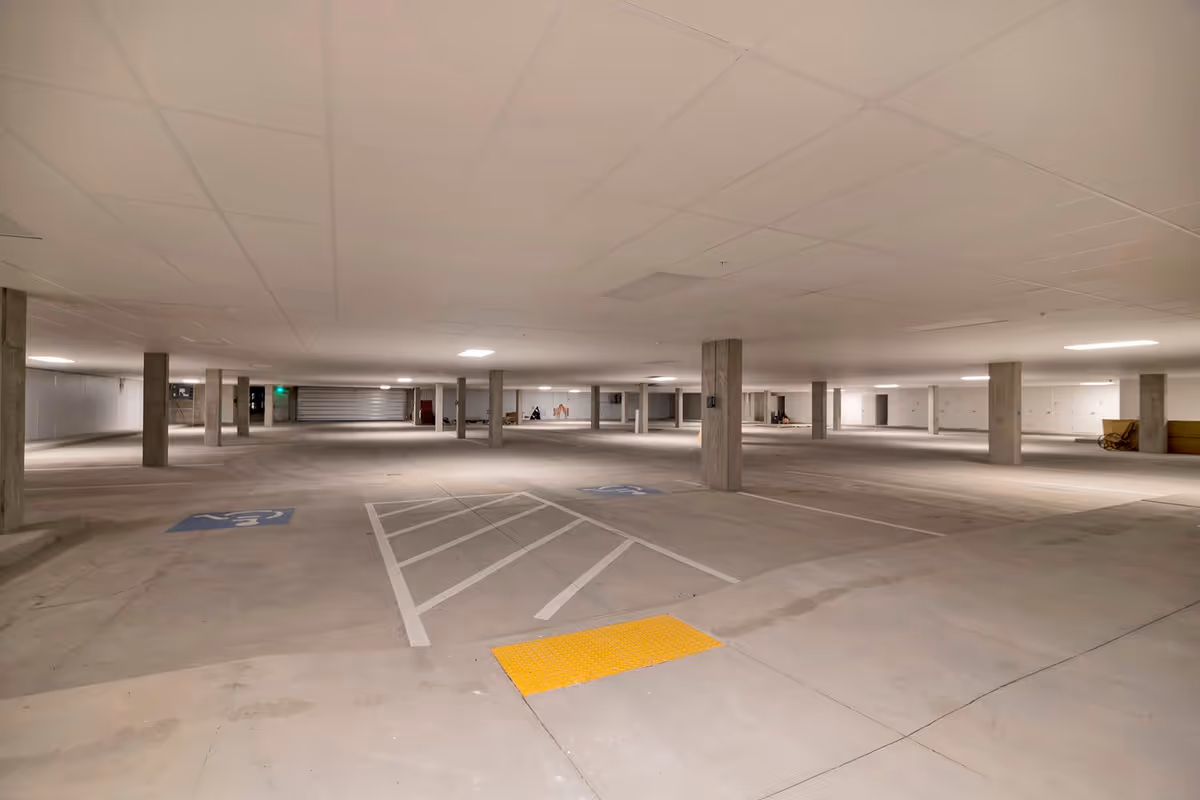 A spacious, empty indoor parking garage with concrete floors, multiple concrete support columns, and overhead fluorescent lighting. There are marked parking spaces including handicapped spots and a yellow tactile paving area near the foreground.