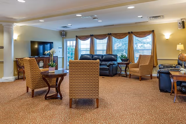 A spacious living room area in a senior living facility with patterned carpet, a black leather sofa, two upholstered armchairs, a wooden TV stand with a flat-screen television, side tables with lamps and plants, large windows with gold curtains, and soft lighting from wall sconces and ceiling lights.