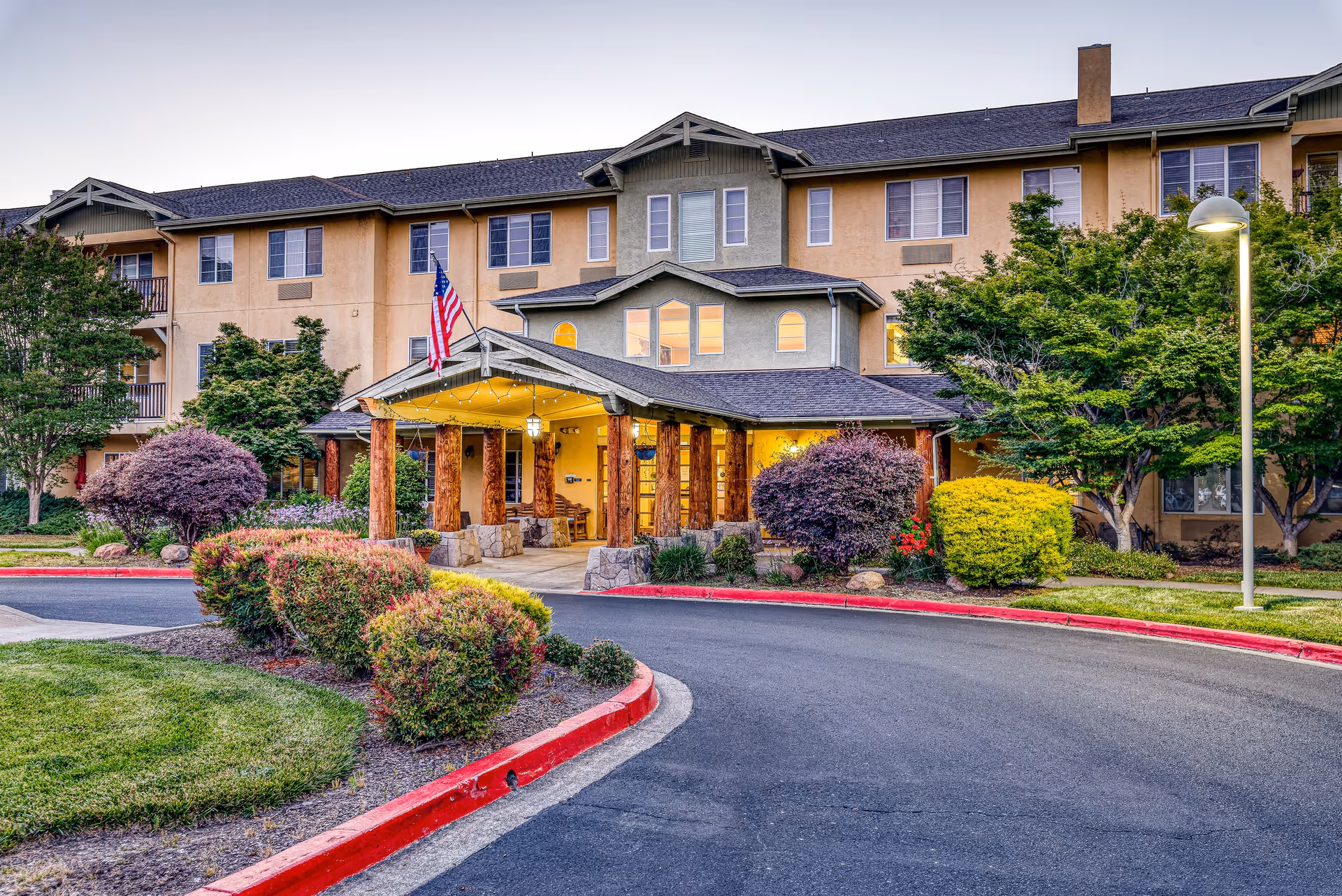 Exterior view of Solstice Senior Living at Santa Rosa building entrance with a covered porch supported by wooden pillars, surrounded by landscaped bushes and trees, and an American flag displayed near the entrance.