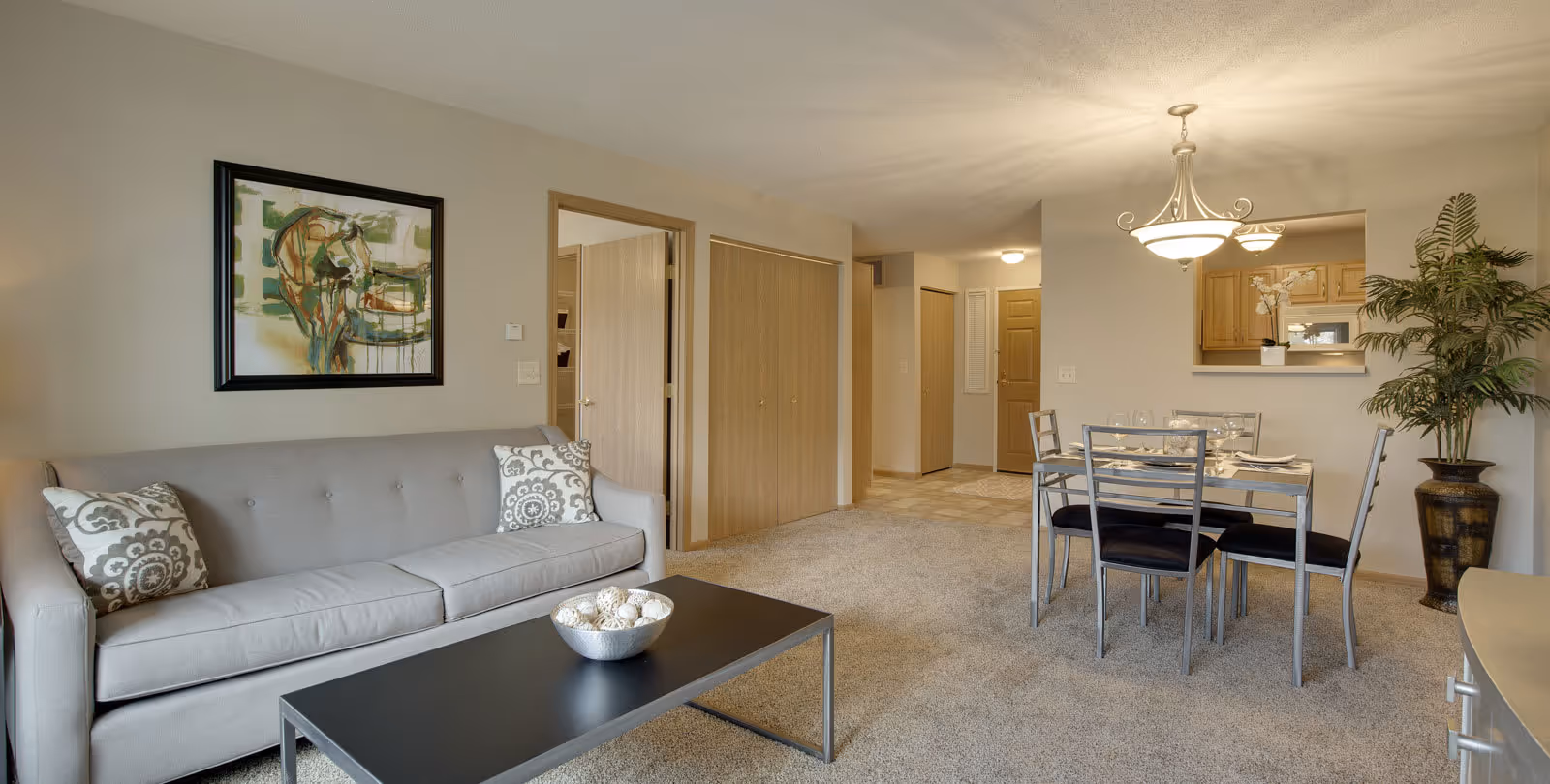 A living and dining area in a senior living facility with a light gray sofa adorned with patterned pillows, a black coffee table with a decorative bowl, a dining table set for four with black cushioned chairs, a large potted plant in the corner, and a kitchen visible through a pass-through window. The walls are beige, and there is a framed abstract painting above the sofa.