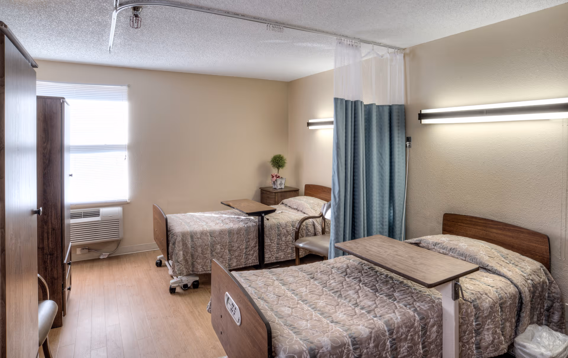 A shared bedroom in a healthcare and rehabilitation center with two hospital-style beds, each with a beige patterned bedspread and an overbed table. There is a privacy curtain between the beds, a wooden nightstand with a small plant and flowers, a chair, a window with blinds, and a wooden wardrobe. The room has light-colored walls and wood flooring.