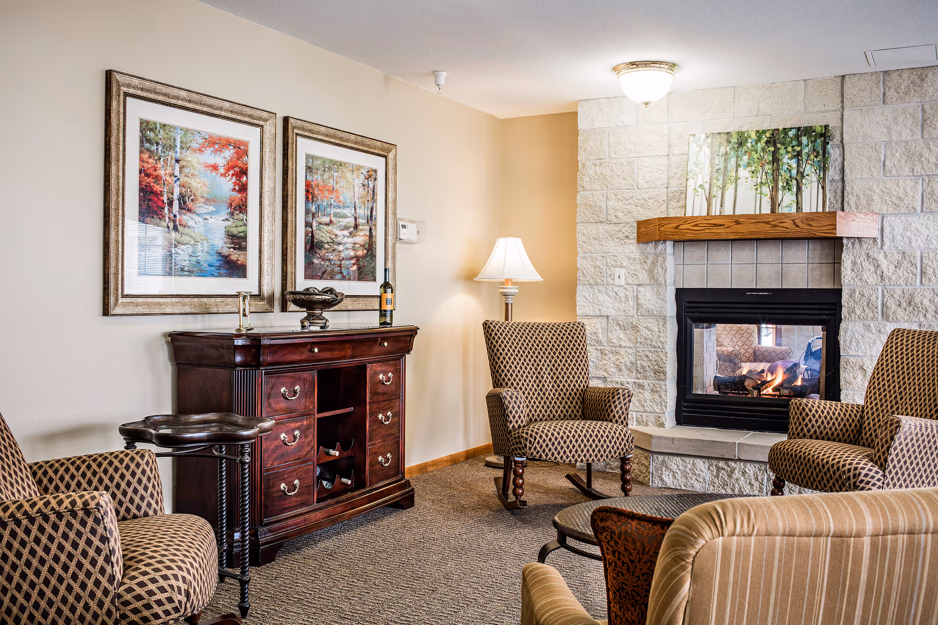 Cozy living room with patterned armchairs arranged around a lit fireplace, a wooden cabinet, framed artwork, and a table lamp.