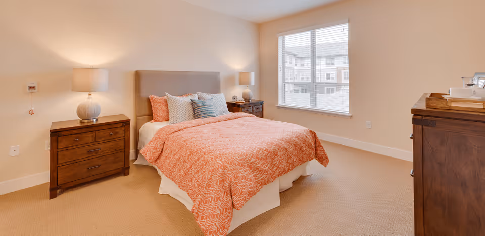 A bright and neatly arranged bedroom with a double bed covered in an orange patterned bedspread and multiple pillows. The room features two wooden nightstands with matching lamps on either side of the bed, a window with blinds letting in natural light, and a wooden dresser with a tray on top.