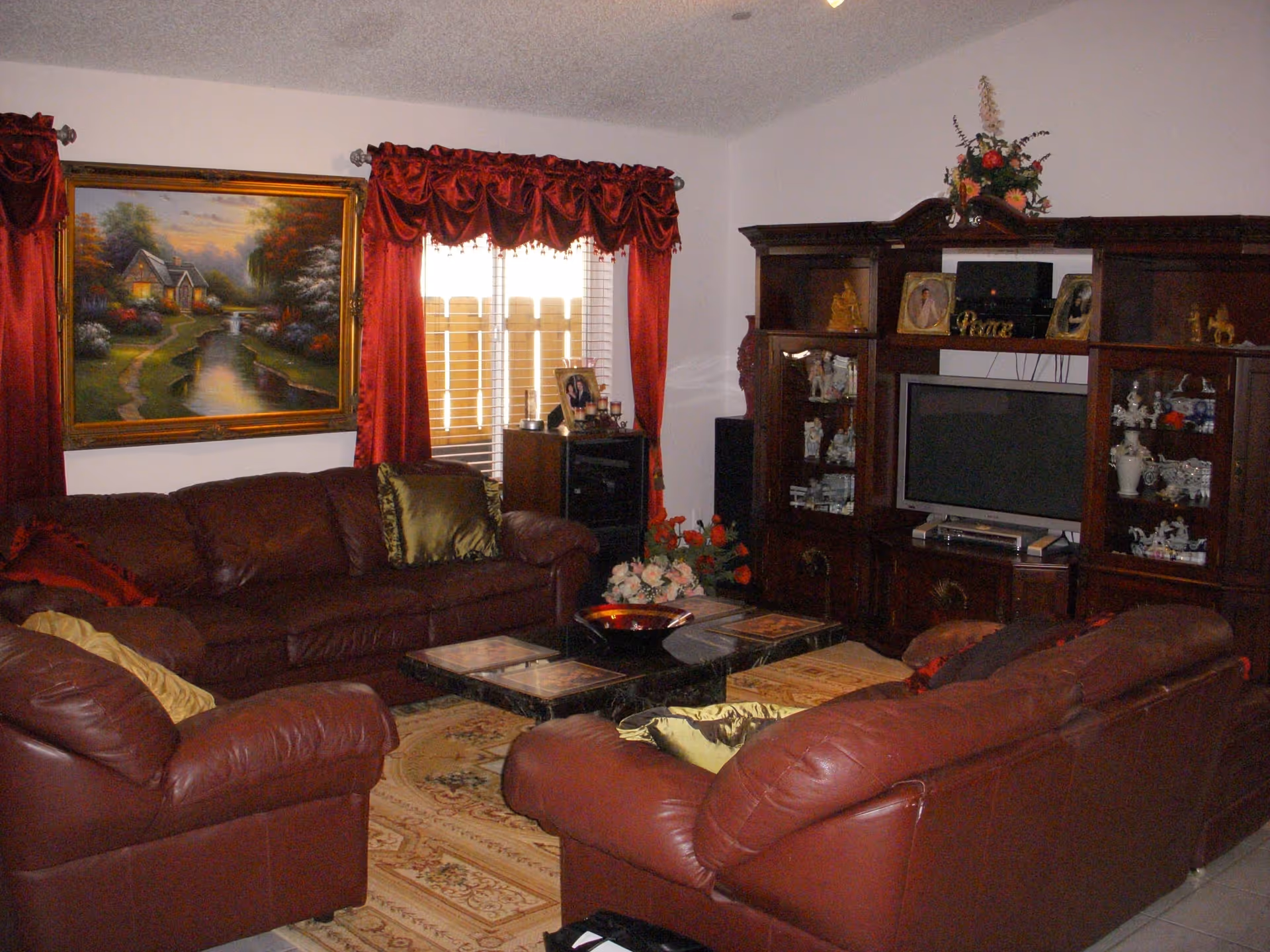 Cozy living room with burgundy leather sofas, a wooden entertainment center with a TV, a coffee table and a framed landscape painting by a window with red curtains.