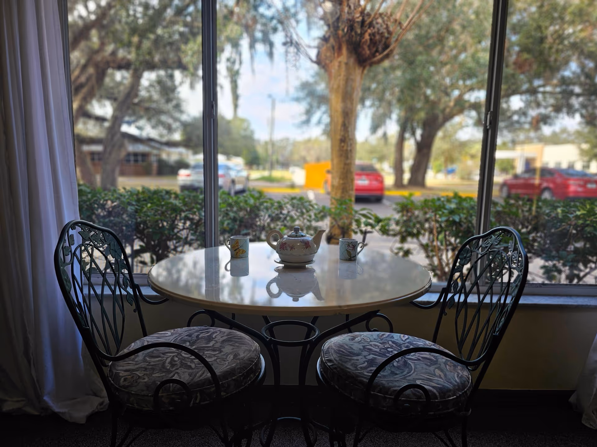 A small round table with a teapot and two cups on it, positioned in front of a large window. Two cushioned metal chairs are placed on either side of the table. Outside the window, there are bushes, trees, and parked cars visible.