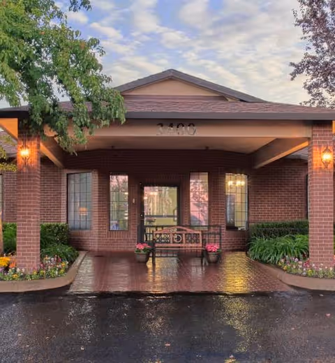 Covered brick entrance with columns, a bench and potted flowers under a lit portico.