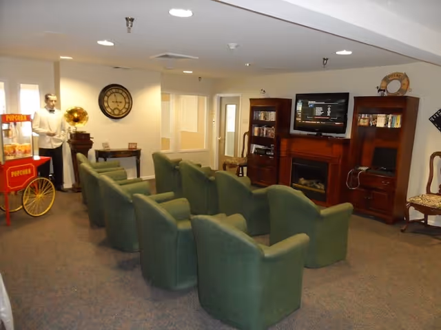 A cozy common area with two rows of green upholstered chairs facing a wall-mounted TV above a wooden fireplace. On either side of the fireplace are wooden cabinets filled with books and decorative items. To the left, there is a vintage-style popcorn machine and a mannequin dressed as a waiter standing next to a gramophone. The room has beige walls and carpeted flooring.