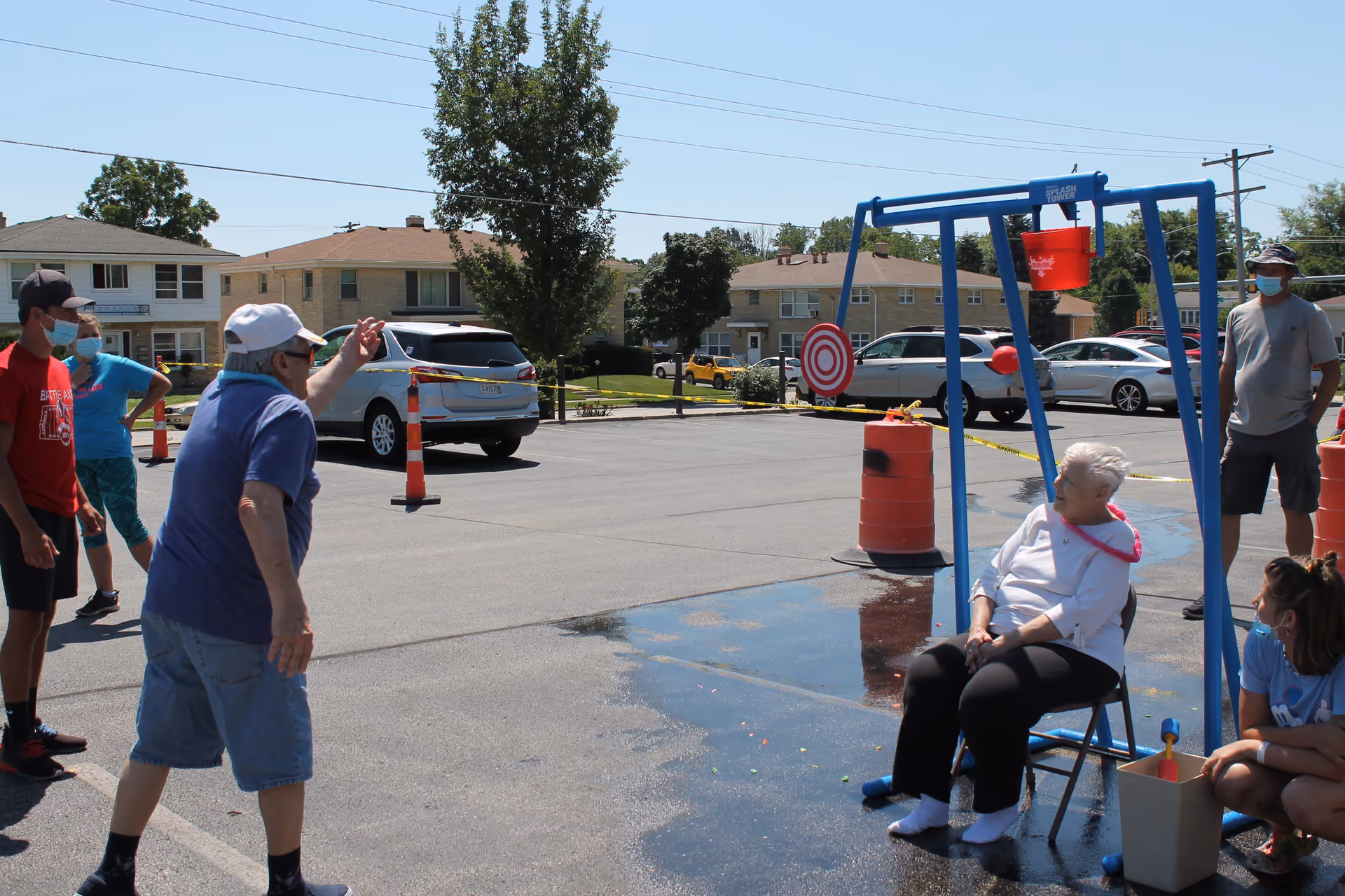An elderly woman sits on a chair under a blue frame with a red bucket above her head in a parking lot. A man wearing a blue shirt and shorts is throwing a ball towards the bucket. Several people, some wearing masks, watch the activity. There are cars and houses in the background under a clear blue sky.