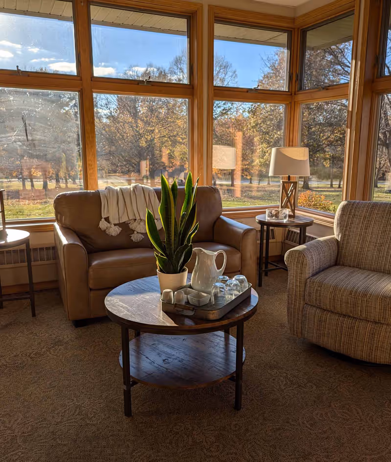 Sunlit sitting area with a leather loveseat, upholstered armchair, round coffee table holding a plant and pitcher, and large windows showing autumn trees.