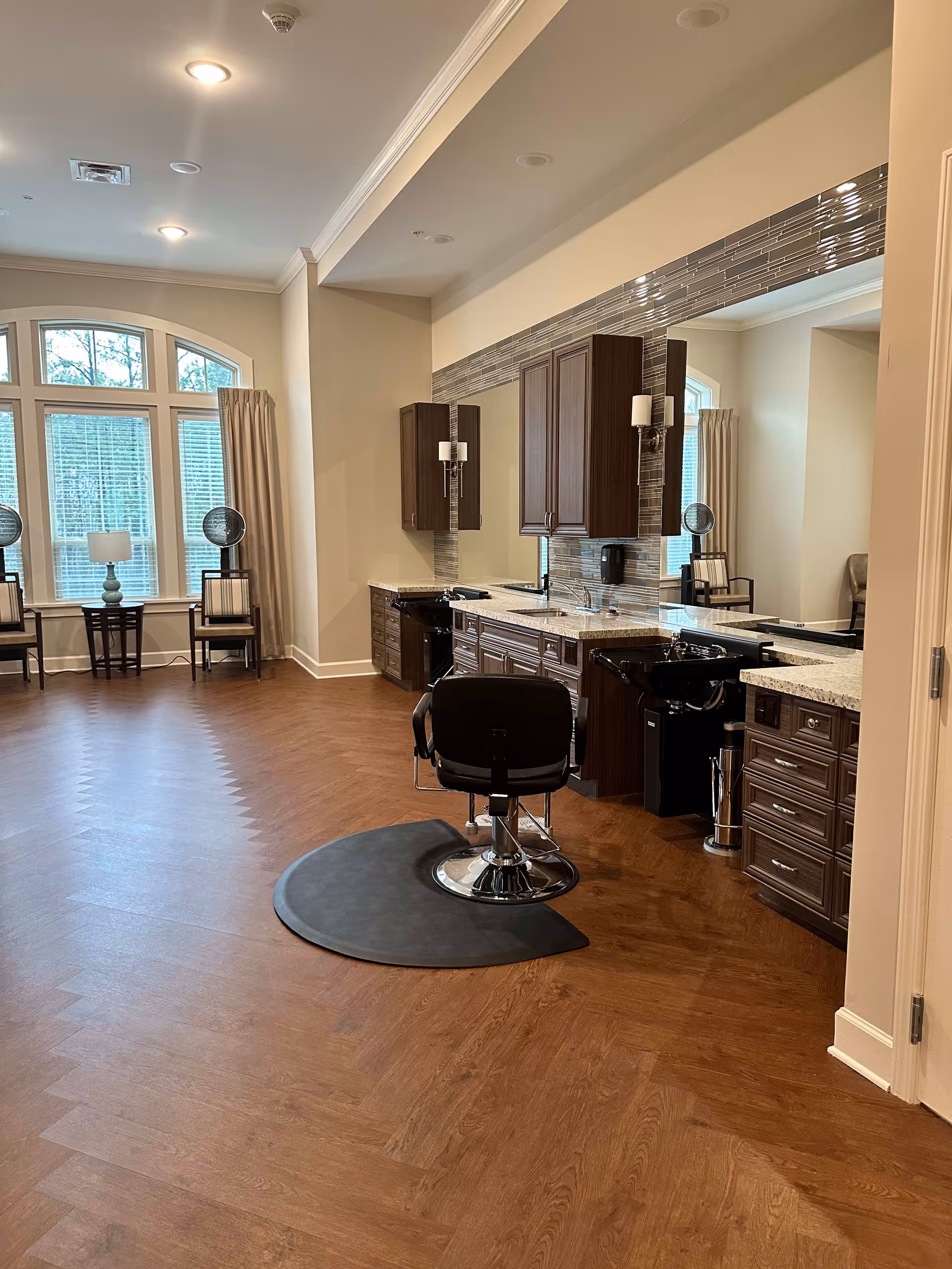 Interior view of a salon area in a senior living facility with a single black salon chair on a protective mat in front of a large mirror and wooden cabinetry. The room has wood-patterned flooring, beige walls, and large windows with beige curtains letting in natural light. Two chairs and a small table with a lamp are positioned near the windows.