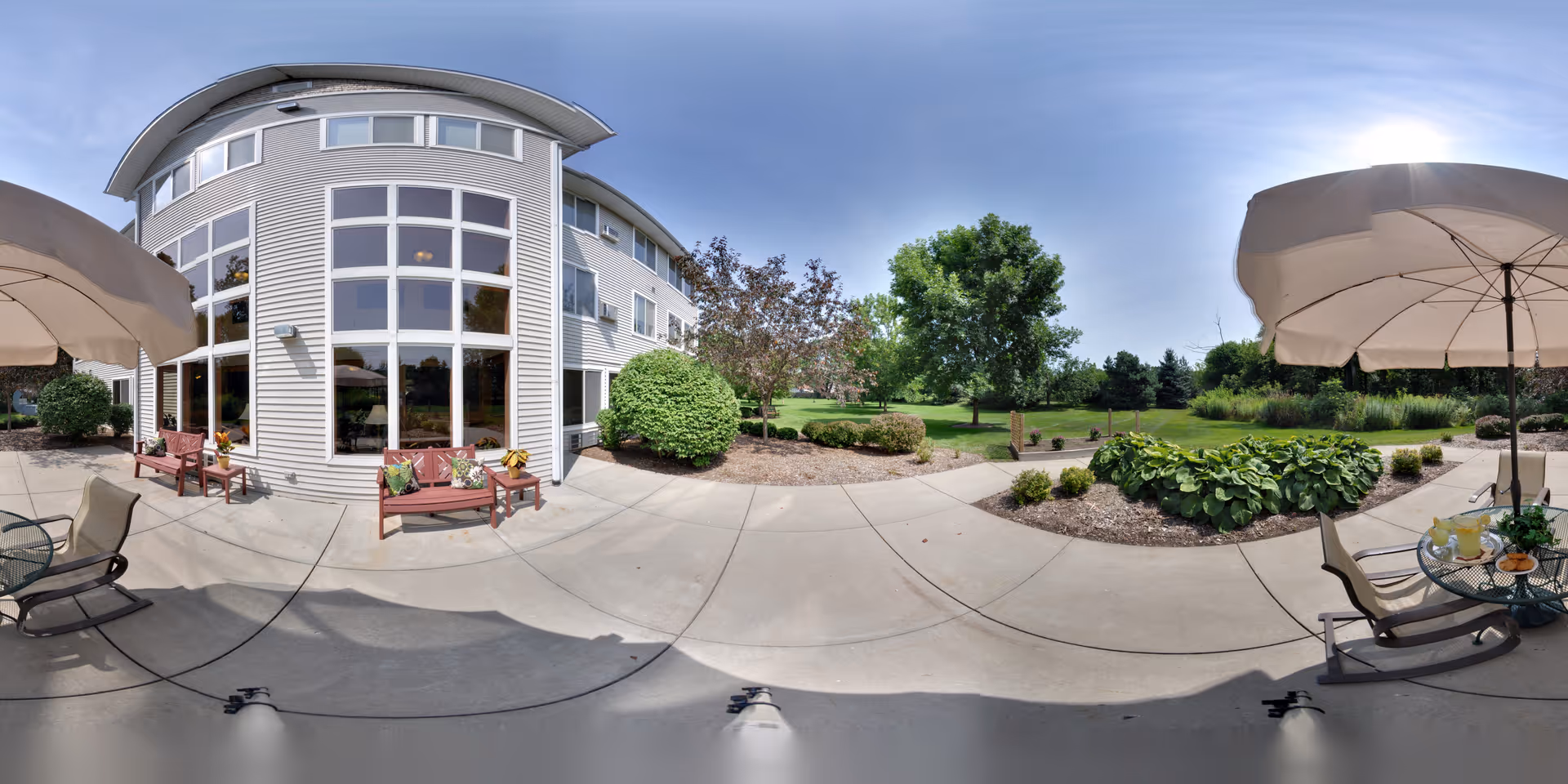 Outdoor patio area at Charter Senior Living of Kenosha featuring concrete walkways, benches with cushions, patio tables with umbrellas, and a landscaped garden with bushes and trees under a clear blue sky.