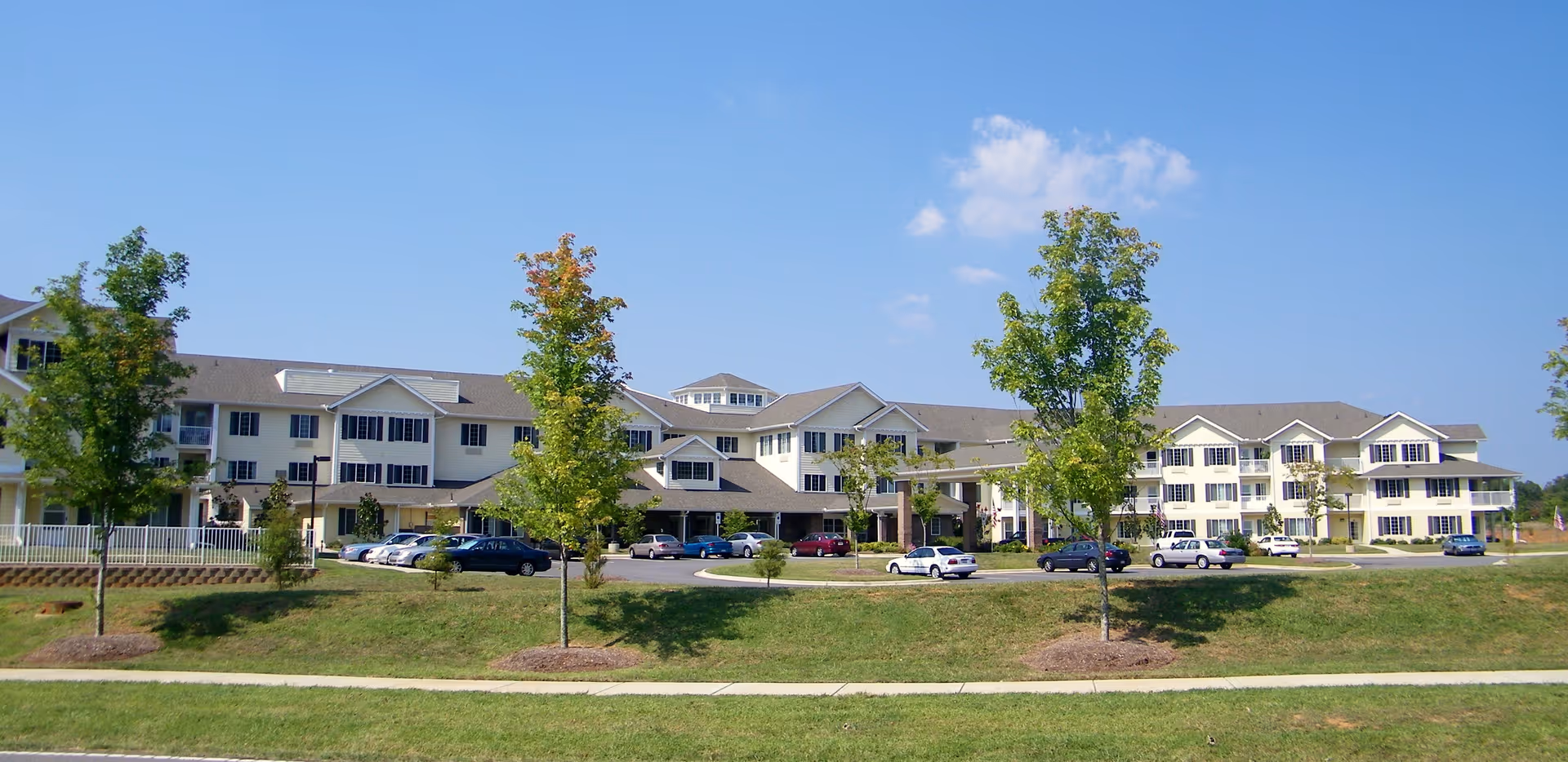 Wide exterior view of a large, multi-story retirement facility building with several windows and a parking lot in front. There are a few trees and a grassy area between the building and the sidewalk under a clear blue sky.