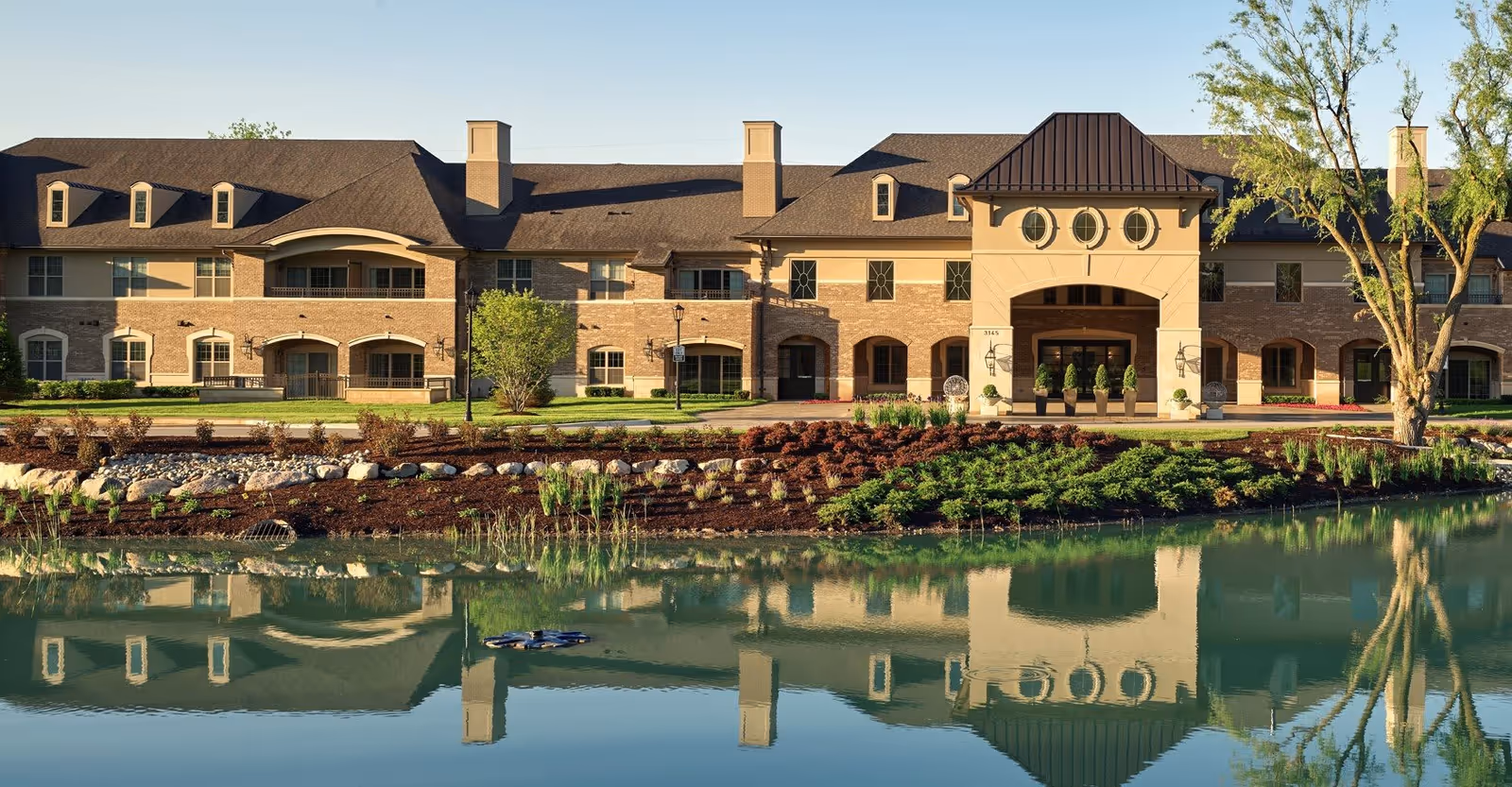 Exterior view of a large senior living facility building with a pond in the foreground reflecting the building and surrounding landscaping. The building has a combination of brick and light-colored walls with multiple windows, a covered entrance with three circular windows above it, and a well-maintained garden area with shrubs and trees.
