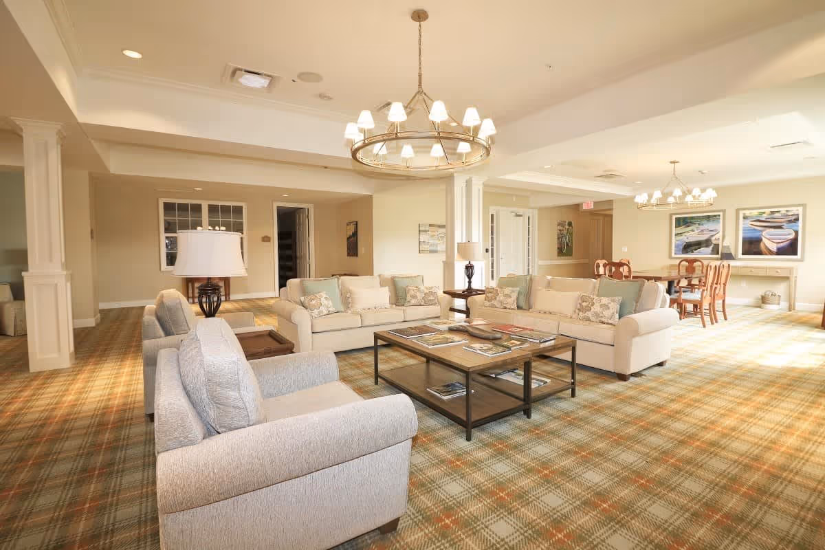 A spacious senior living common area with beige sofas, armchairs, a wooden coffee table with magazines, and two chandeliers hanging from the ceiling. The room has a plaid carpet, light-colored walls, and framed artwork on the far wall. There is also a dining table with chairs in the background.