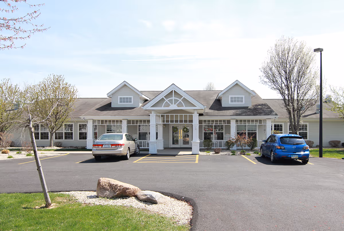 Front exterior view of Campbell Place, a single-story building with a covered entrance supported by white columns. There are two cars parked in front, a silver sedan and a blue hatchback. Trees and landscaping with rocks and grass surround the parking area.