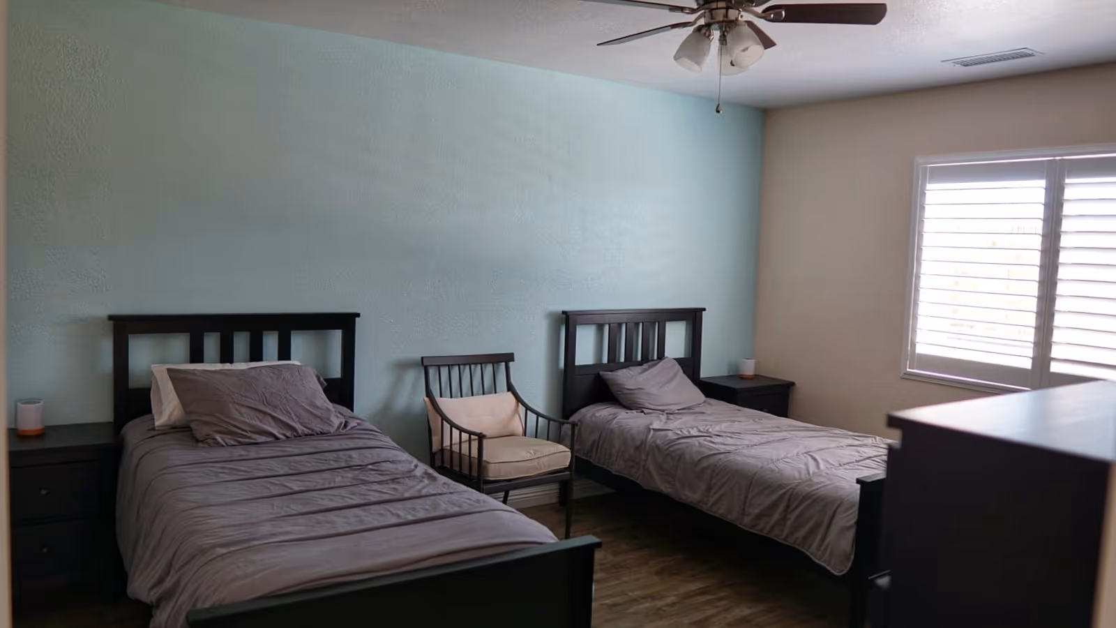 A bedroom with two single beds featuring dark wooden headboards and gray bedding. Between the beds is a wooden chair with a beige cushion. Each bed has a dark wooden nightstand with a small lamp. The room has light blue and beige walls, a ceiling fan with lights, and a window with white blinds letting in natural light.