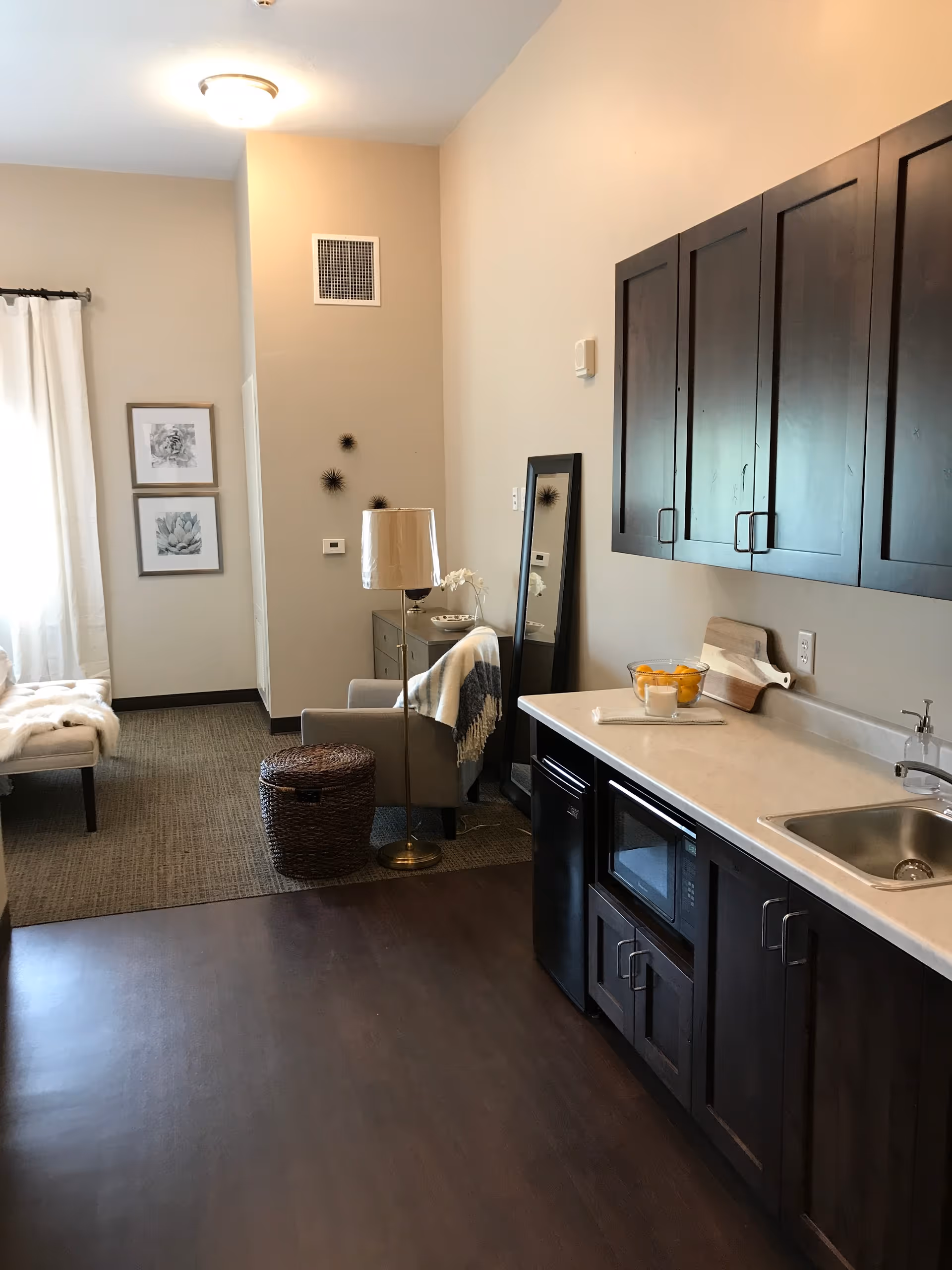 A cozy interior space featuring a kitchenette with dark wood cabinets, a microwave, a small refrigerator, and a sink. To the left, there is a sitting area with a gray armchair draped with a blanket, a floor lamp, a wicker basket, and a tall mirror leaning against the wall. The room has beige walls, framed artwork, and a window with white curtains allowing natural light to enter.