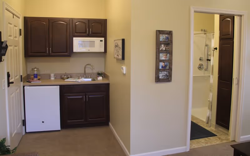 Small kitchenette area with dark wood cabinets, a white microwave, a sink, and a mini refrigerator. To the right, there is an open doorway leading to a bathroom with a shower and a dark wood door. The walls are painted light beige, and there is a photo frame with multiple pictures hanging on the wall between the kitchenette and bathroom.