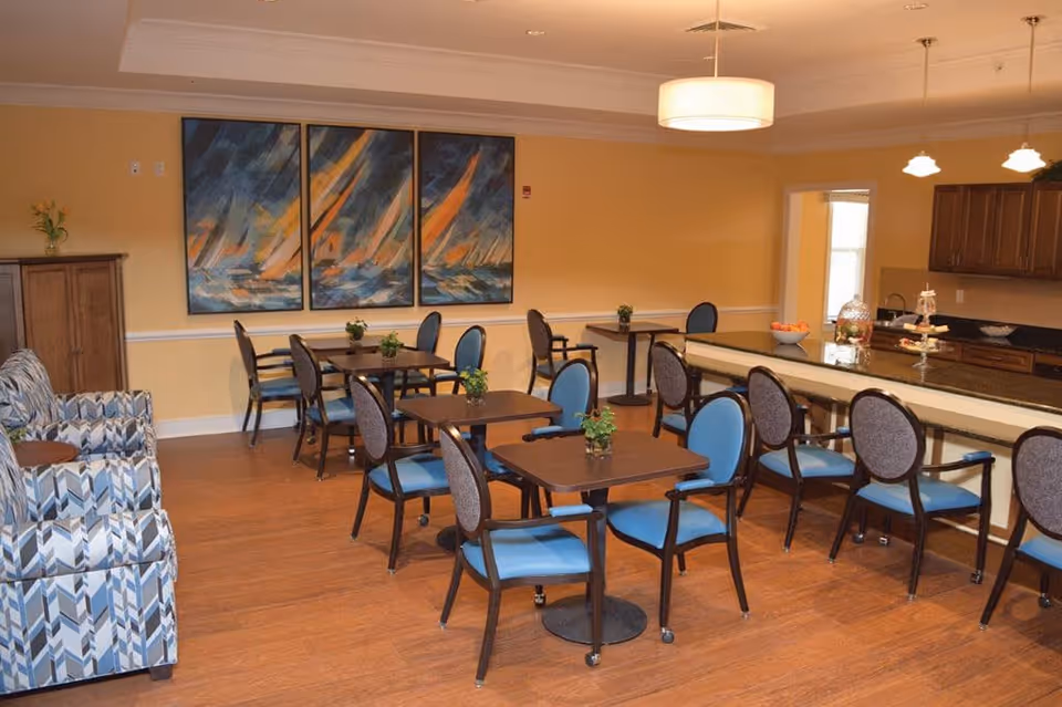 A dining area in a senior living facility with several square wooden tables and blue cushioned chairs arranged neatly. There is a long counter with additional seating on one side, and a kitchen area with wooden cabinets and pendant lights in the background. The walls are painted yellow and decorated with a three-panel abstract painting. The floor is wooden, and there are a few small plants on the tables.