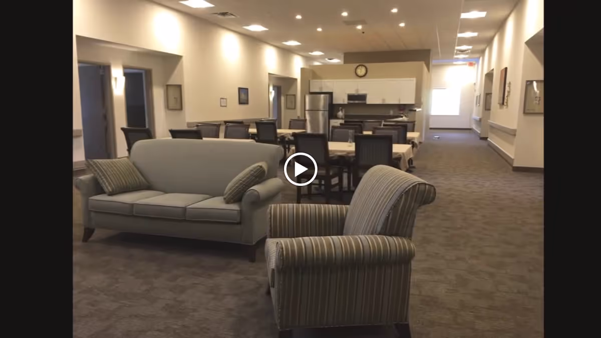 Communal senior living lounge and dining area with a sofa and armchair in the foreground and dining tables and a kitchenette in the background.