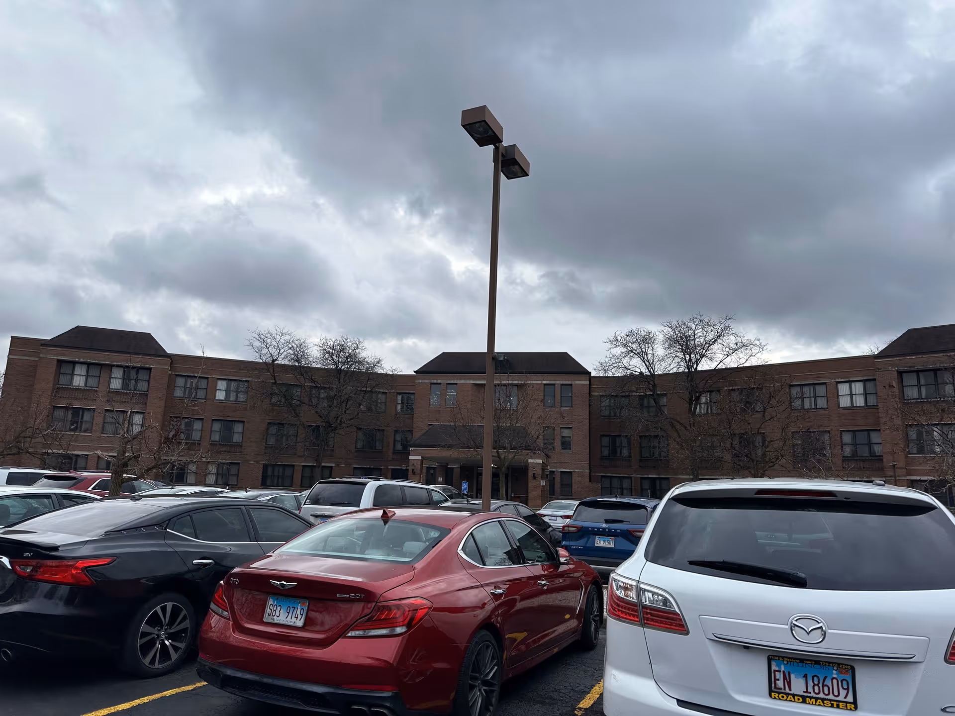 Exterior view of a three-story brick building with multiple windows, identified as Belhaven Nursing Home, under a cloudy sky. In the foreground, there is a parking lot with several parked cars and a tall street lamp in the center.